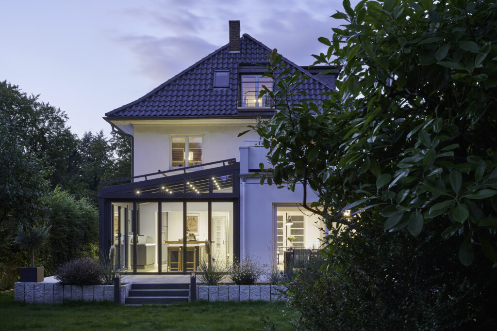 Modernisierter Altbau mit stilvollem Wintergarten aus Glas und Aluminium; abendliche Beleuchtung setzt den überdachten Essbereich im Garten elegant in Szene.